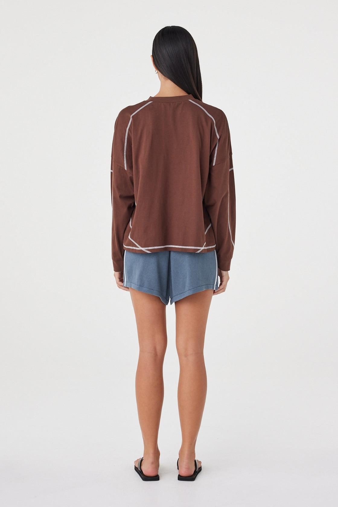 Model wearing the espresso long-sleeve tee with blue shorts and black sandals, showing the dropped shoulders, contrast cream overlock seams, ribbed cuffs and relaxed silhouette in a full-length back view.
