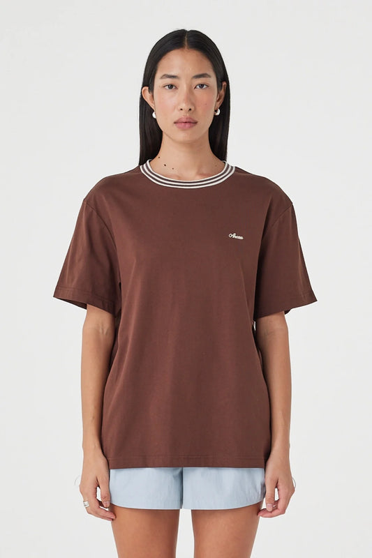Close-up front view of the espresso oversized tee showing the cream contrast neckline and chest logo, styled with pale blue shorts against a clean white studio background.