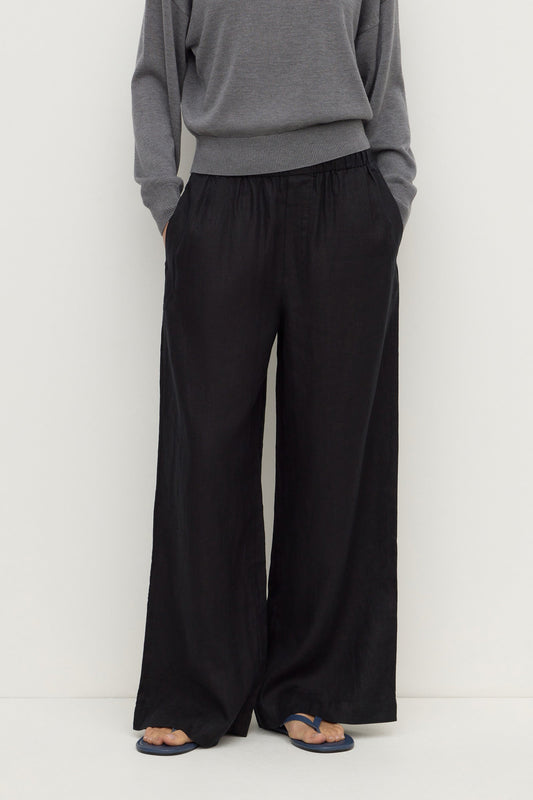 Close-up front view of black wide-leg pants showing the gathered elasticated waistband, side pockets and flowing leg shape.