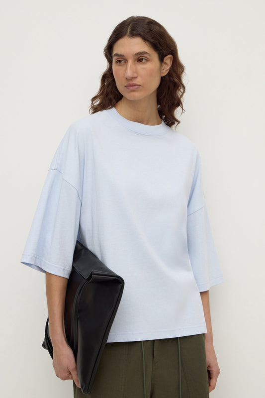 Model wearing a powder blue oversized short-sleeve tee with olive trousers and holding a black clutch, showing the loose silhouette, front view