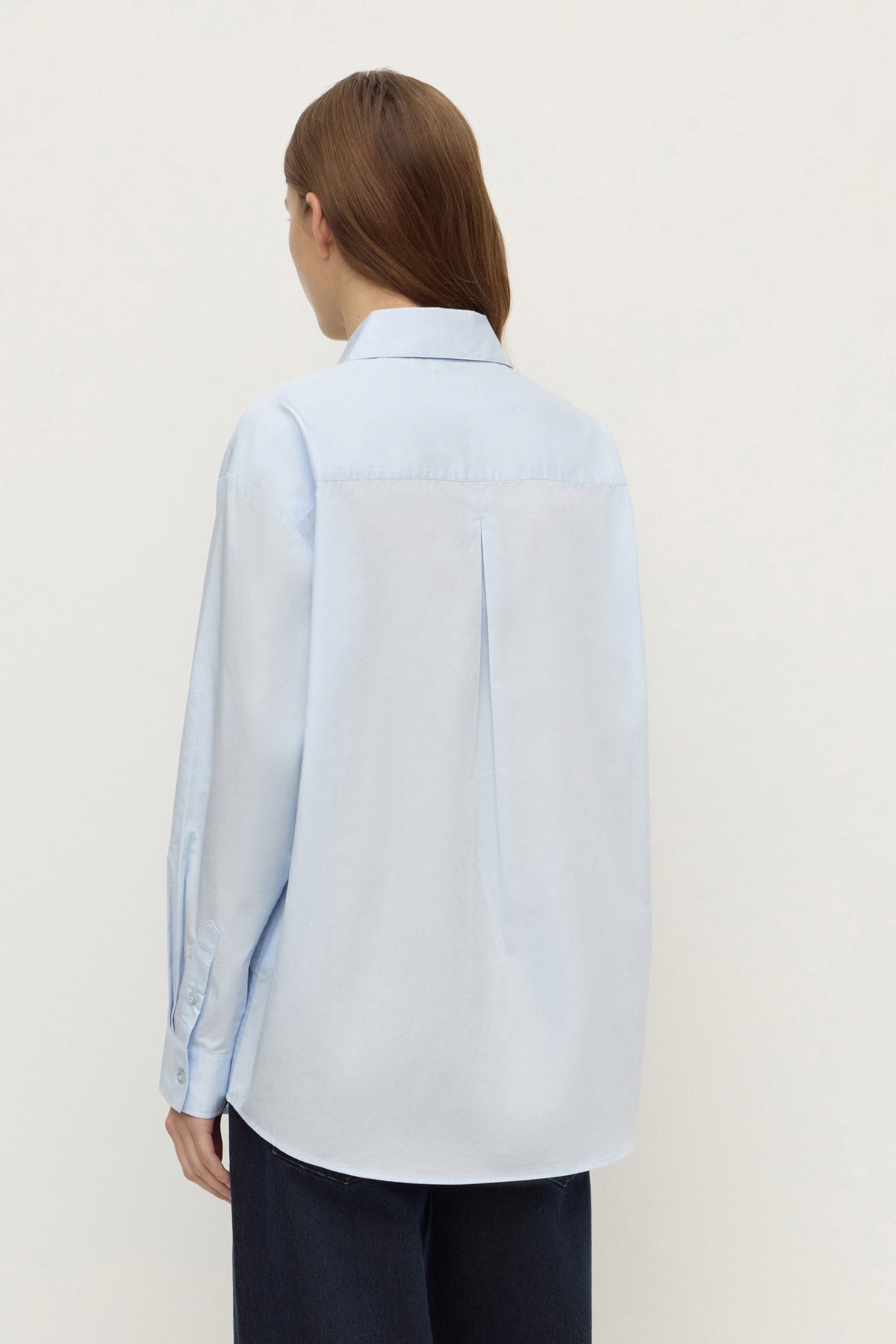 Model wearing a powder blue long-sleeve button-up shirt with navy trousers, showing the back yoke, relaxed silhouette and curved hem, back view