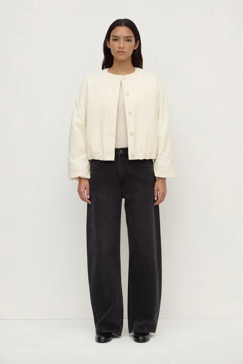 A model stands against a plain white studio background wearing a cream-colored cropped button-up jacket over a light beige top and black wide-leg trousers.
