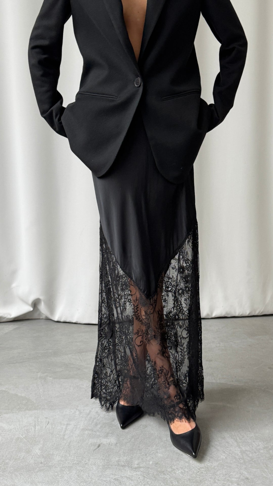 A front view of the black skirt styled with a tailored blazer, highlighting the satin overlay and transparent floral lace hem.