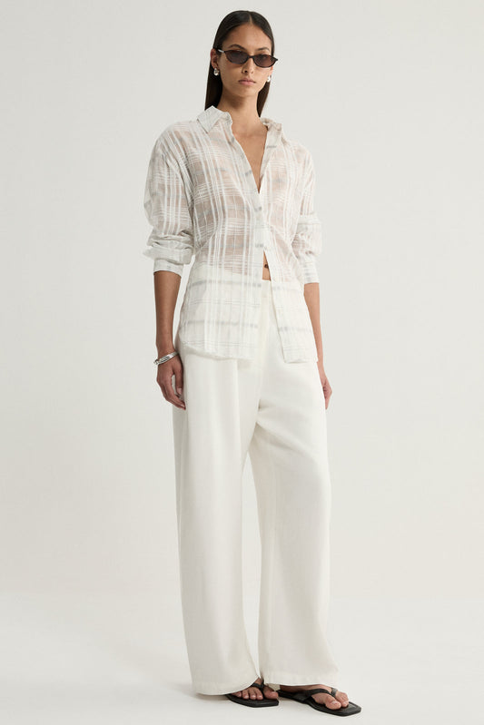 A model wearing a white sheer check shirt with neutral vertical and horizontal lines, a collared neckline and long sleeves, paired with ivory wide-leg trousers and black flat sandals in a full-length front view.
