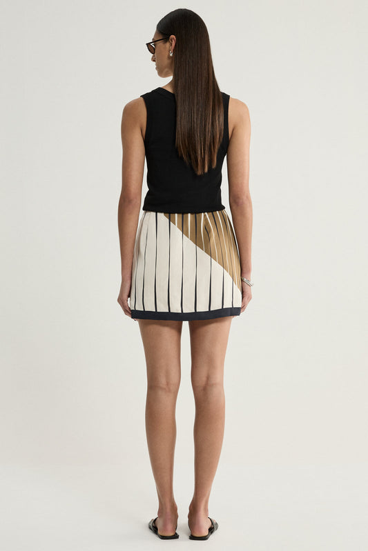 A rear view of a model wearing the cream striped mini skirt with a diagonal moss-coloured panel across the back, fine dark vertical lines and a contrasting dark hem, styled with a black sleeveless top and black flip-flops.