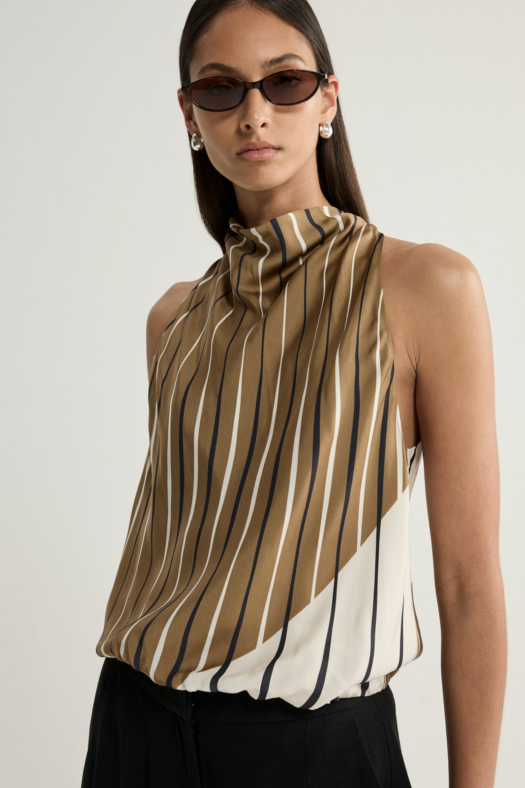 A close-up portrait of a model wearing a moss, cream and dark striped top, highlighting the gathered high neckline, fluid fabric, curved colour-blocked pattern, narrow dark sunglasses and silver earrings.