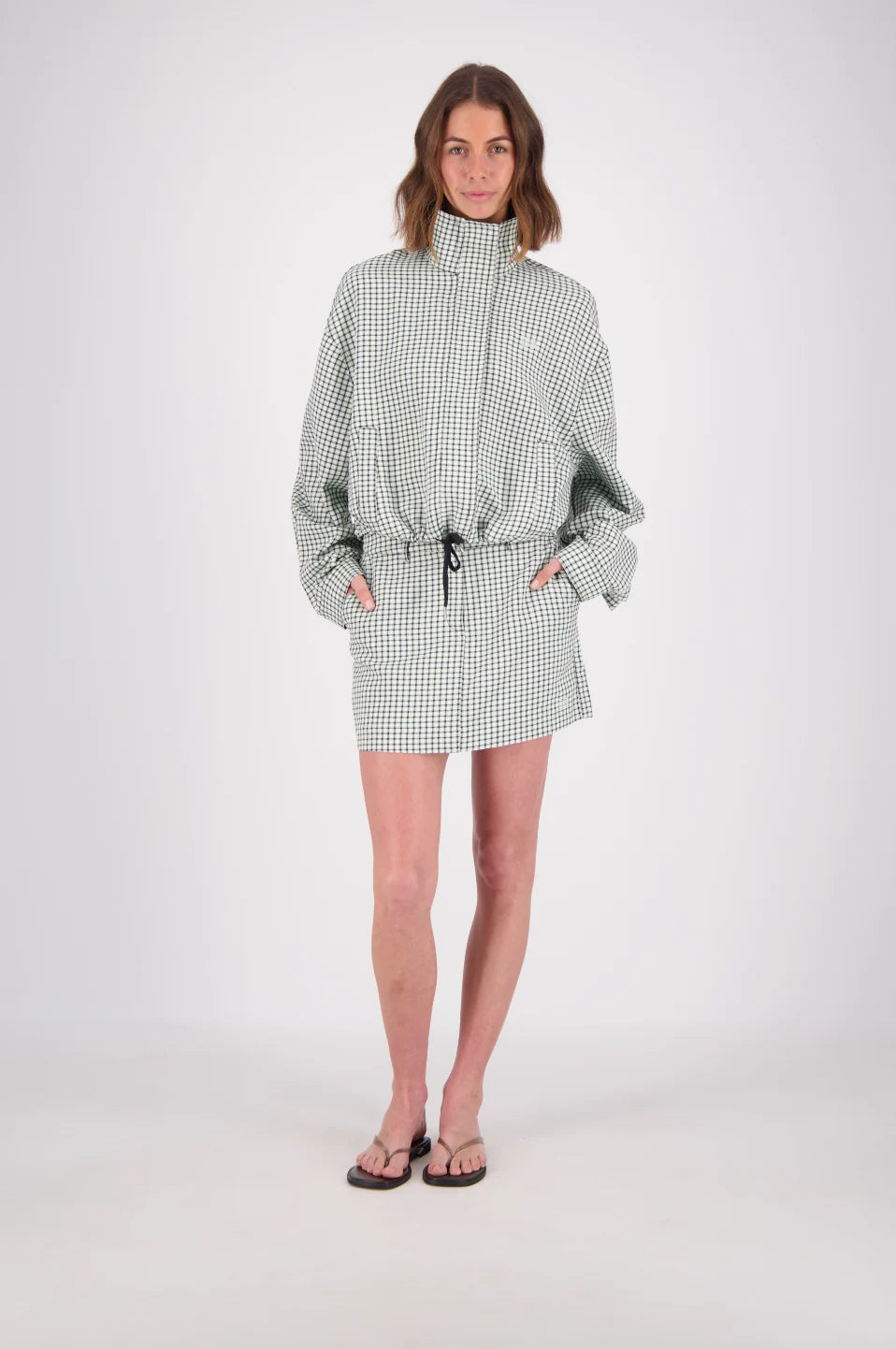 A front-facing studio view of a white and mint check bomber jacket worn with a matching mini skirt, featuring a high stand collar, long sleeves, side pockets and a black drawstring hem.