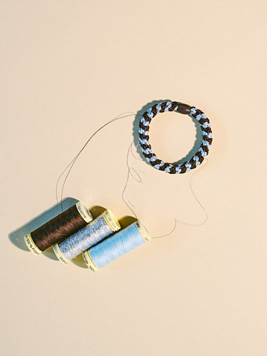 Brown and sea blue woven hair tie displayed beside matching thread spools on a neutral background.