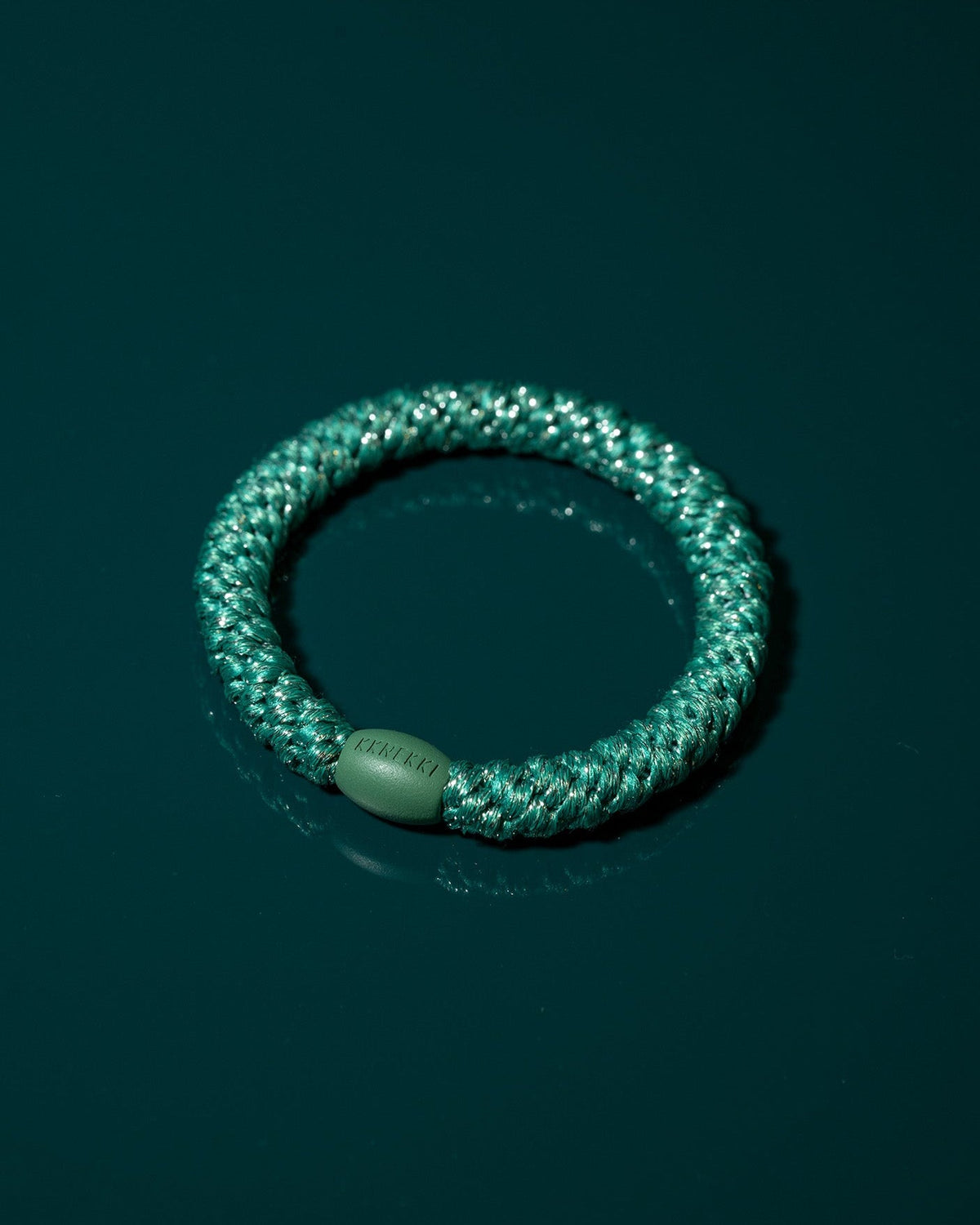 Close-up product shot of the teal glitter hair tie resting on a deep green surface, highlighting its woven texture, metallic sparkle and rounded bead.