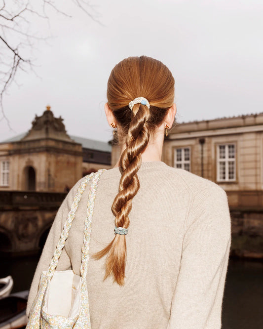 Slim teal and gold glitter hair tie securing a woman's braided ponytail outdoors in front of a historic building.