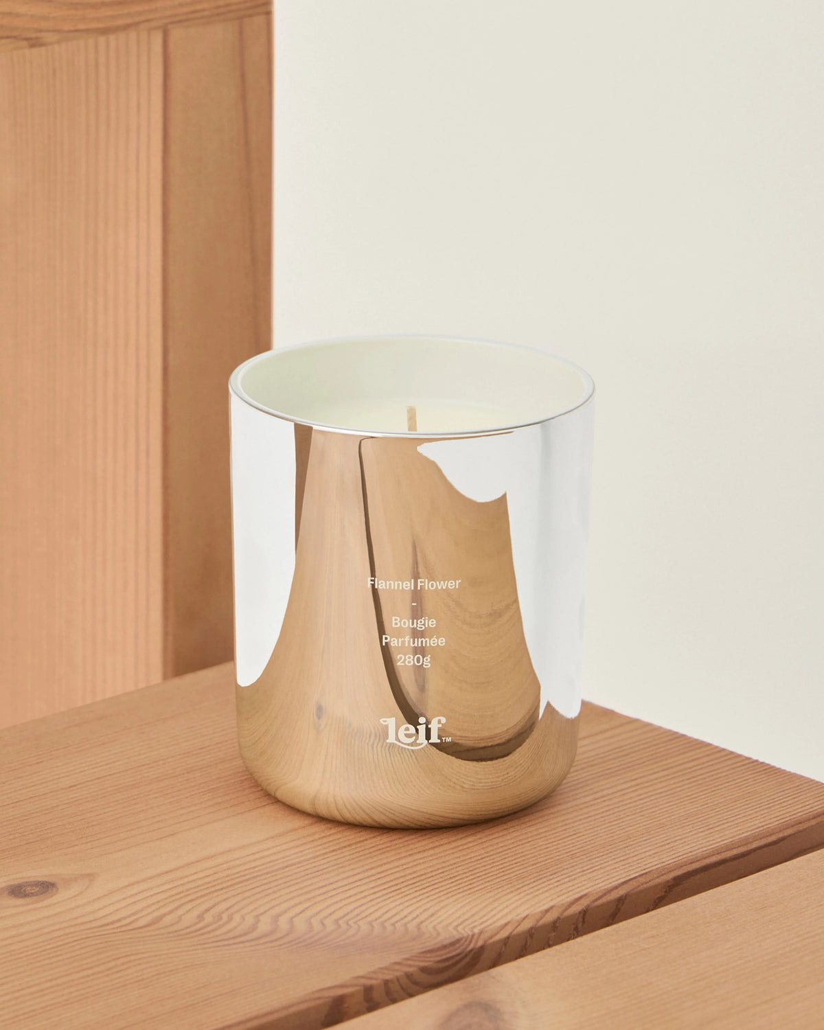 A scented candle in a reflective chrome glass vessel sits on a wooden shelf, showing its open top and pale wax.
