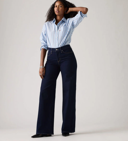 Model wearing the dark indigo wide-leg jeans with a blue striped shirt and black pointed shoes, showing the high-rise waist, fitted hip and full-length wide legs in a full-length front view.