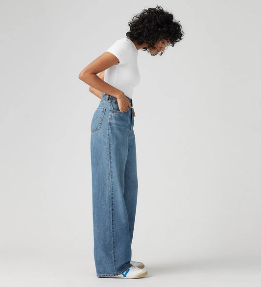 Model wearing light blue high-rise wide-leg jeans with a white T-shirt and sneakers, showing the high waist, side pockets and straight wide-leg profile, side view