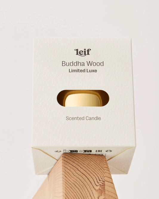 A cream Buddha Wood Limited Luxe candle box with a window revealing the gold vessel is displayed on a wooden block.
