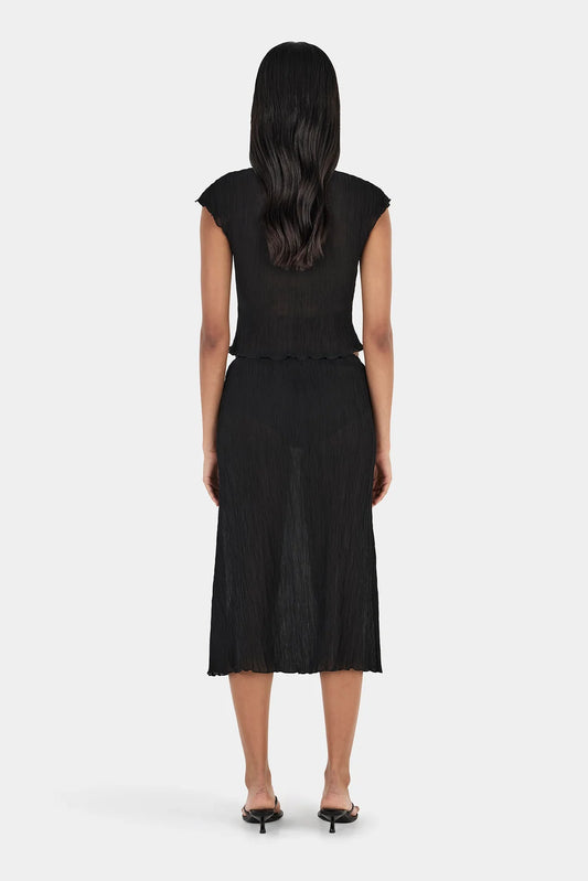 Back view of a model wearing a black midi skirt with a crinkled texture, elasticated waistband, straight silhouette, semi-sheer appearance, and slightly uneven hemline, paired with a black short-sleeve top and black heeled sandals.