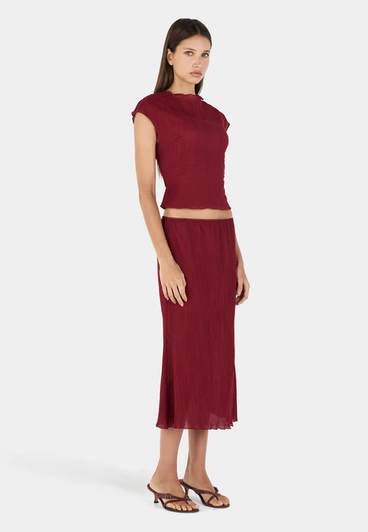 Three-quarter front view of a model wearing a deep crimson midi skirt with a gathered elasticated waistband, fine crinkled texture, straight silhouette, semi-sheer appearance, and below-the-knee hemline, paired with a matching short-sleeve top and brown strappy heeled sandals.