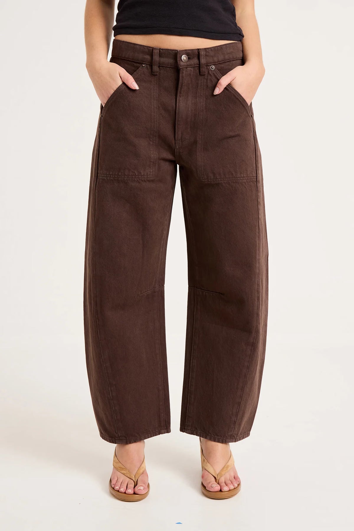 A close-up front view of chocolate-brown pants worn with a black cropped top and tan sandals, focusing on the button fastening, zip fly, belt loops, angled front pockets, metal rivets and horizontal stitched panels across the upper legs.