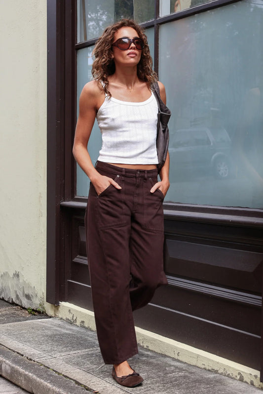 A model with curly brown hair wears chocolate-brown barrel-leg pants with a white cropped camisole, oversized dark sunglasses and black flip-flops, standing on a city pavement beside a large dark-framed shop window. One hand rests in a front pocket while the relaxed legs fall towards the ankles, creating a casual street-style look.