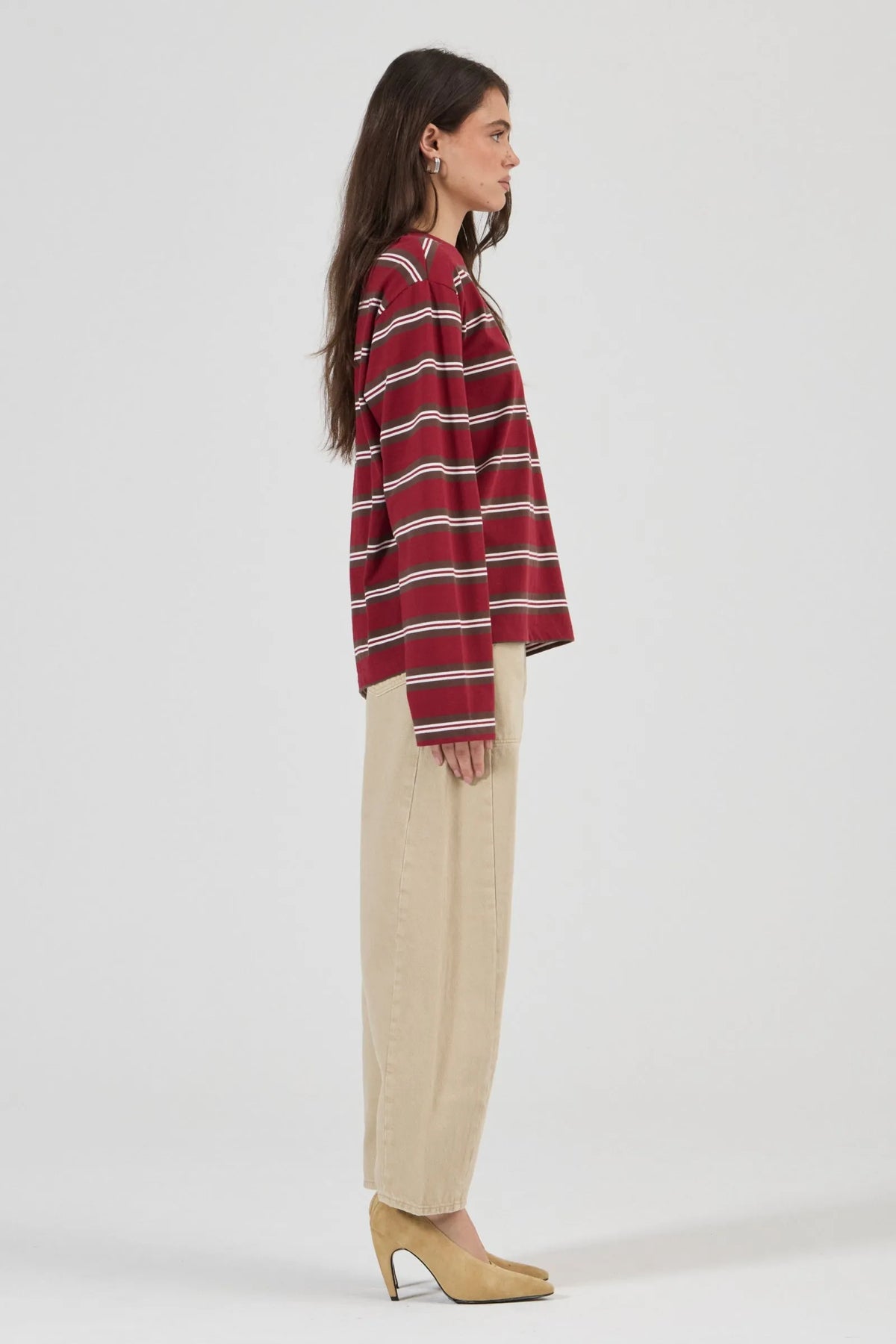 Model wearing the burgundy striped long-sleeve tee with relaxed beige jeans and tan heels, showing the crew neckline, dropped shoulders, long sleeves and relaxed hip-length silhouette in a side view.