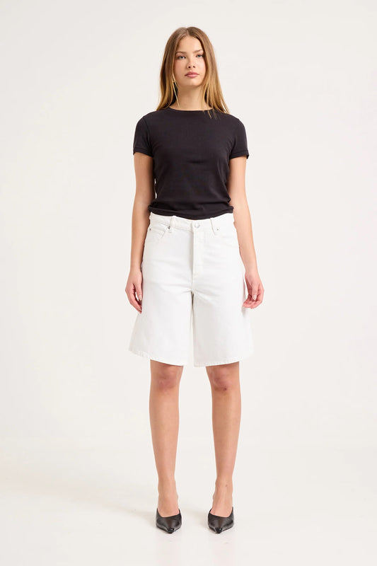 Woman wearing white knee-length denim shorts with a black fitted T-shirt and black pointed shoes in a studio setting.