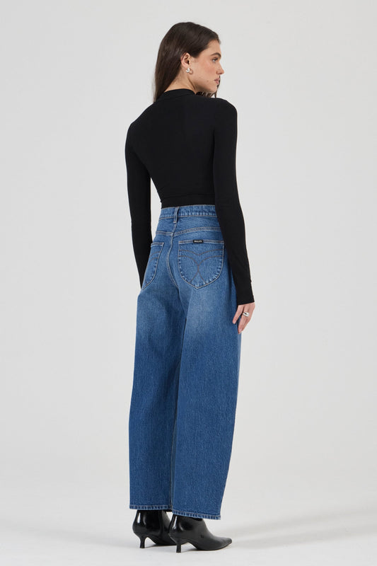 Model wearing the Rolla's Gigi Barrel Jean in Reign with a black turtleneck top, showing the signature angled spade back pockets, full-length back view