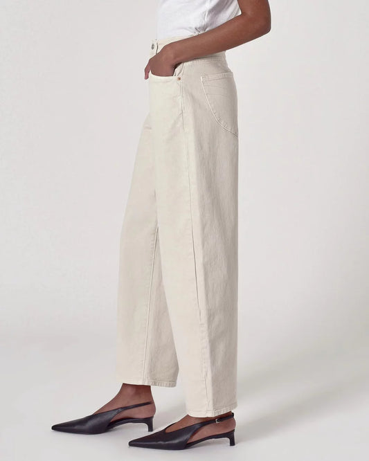 Side view of salt-coloured barrel-leg jeans showing the high-rise waistband, curved leg shape and ankle-length hem, styled with a white top and dark slingback heels.