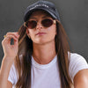 Model wearing the 180EYEWEAR 'Isla' sunglasses in gold with brown lenses and a black cap, close-up lifestyle shot against a charcoal grey background.