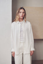 A woman modeling a white long-sleeve button-up shirt and matching white trousers in a minimalist indoor setting with neutral beige and gray walls.