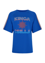 A royal blue short-sleeve t-shirt with 'KINGA CSILLA' printed in a light blue-to-purple gradient on the chest, featuring a purple sunburst graphic in the center. The sleeves have thin orange stripes near the cuffs.