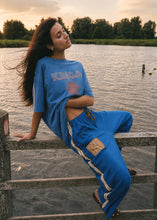 A woman wearing a blue oversized graphic T-shirt and matching blue jogger pants with white side stripes, leaning on a stone railing by a lake at sunset.
