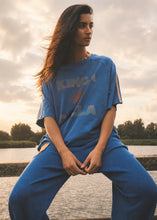 A woman wearing a blue oversized graphic T-shirt and matching blue sweatpants sits outdoors by a body of water at sunset, with the shirt showing a large multicolor 'KING OF LA' graphic.