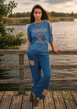 A woman stands on a wooden dock by a lake wearing a matching blue lounge set with a graphic T-shirt and wide-leg pants. The set features a blue T-shirt with 'KINGA ISLA' text and a red graphic, paired with matching blue pants that have brown patch details on the upper thighs and cuffs.