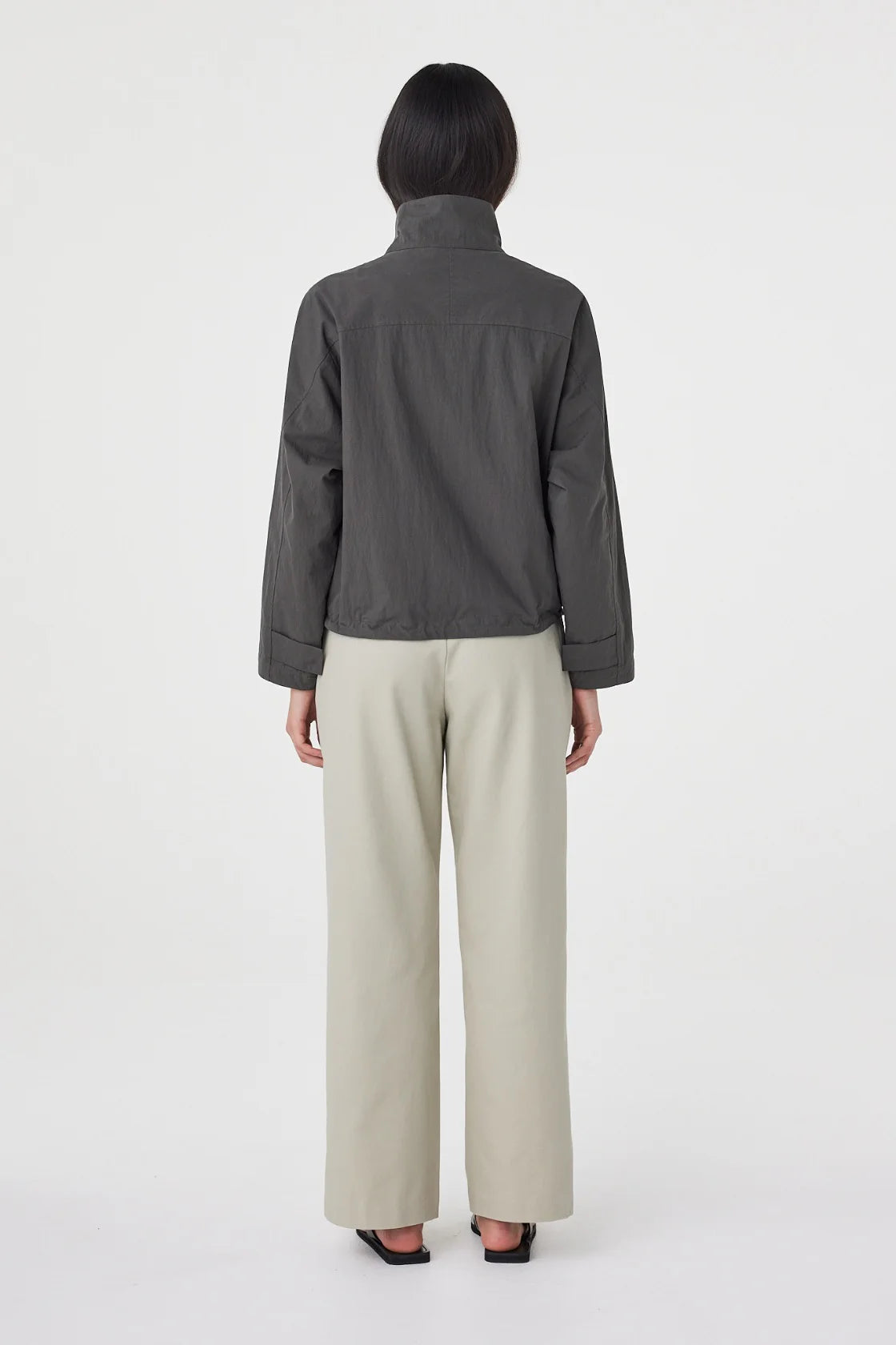 A model wears the charcoal cropped jacket in a back studio view showing the yoke seam and gathered hem.

