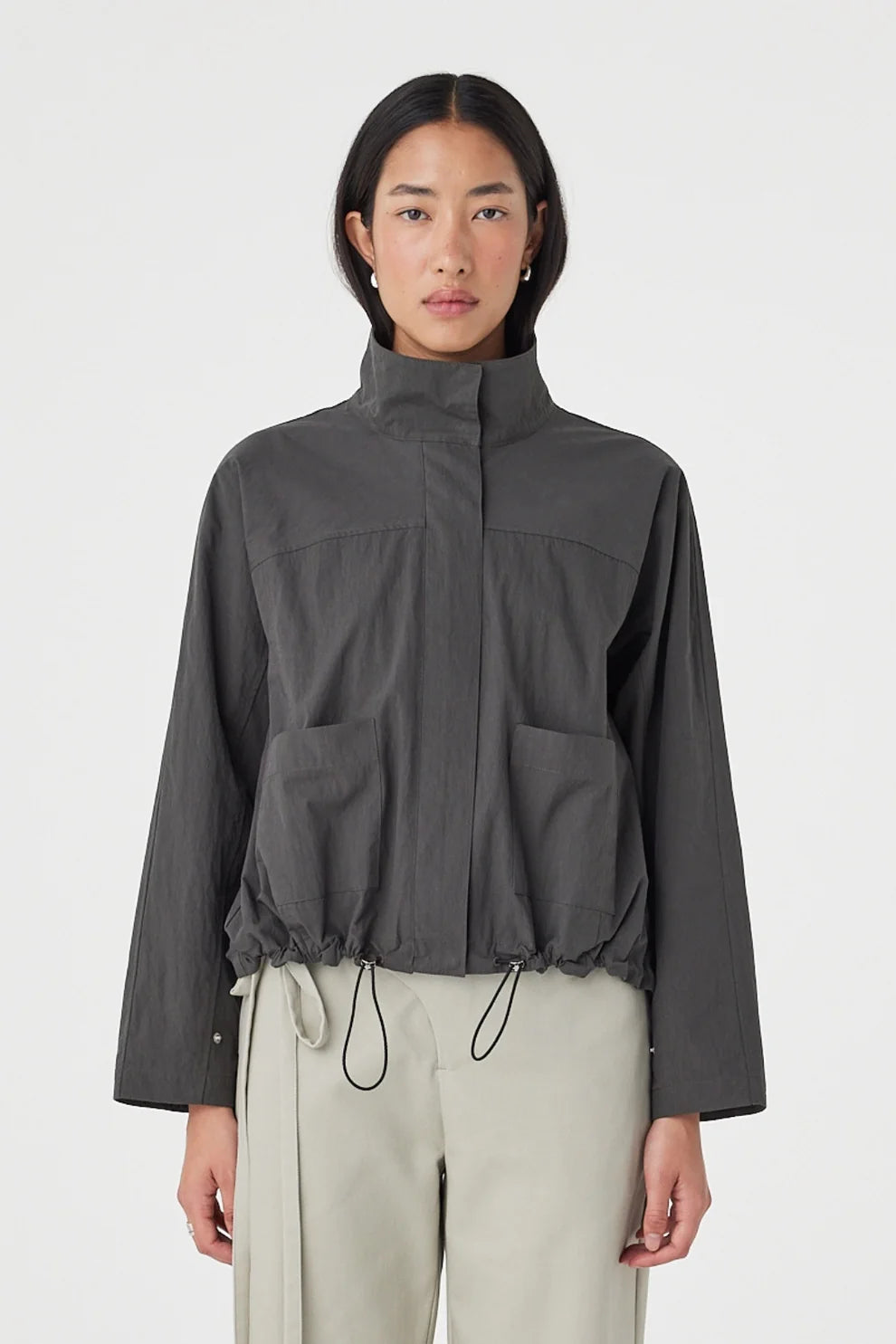 A close-up shows the high collar, concealed front fastening, patch pockets and adjustable drawcord hem on the charcoal jacket.