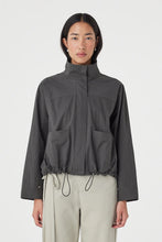 A close-up shows the high collar, concealed front fastening, patch pockets and adjustable drawcord hem on the charcoal jacket.