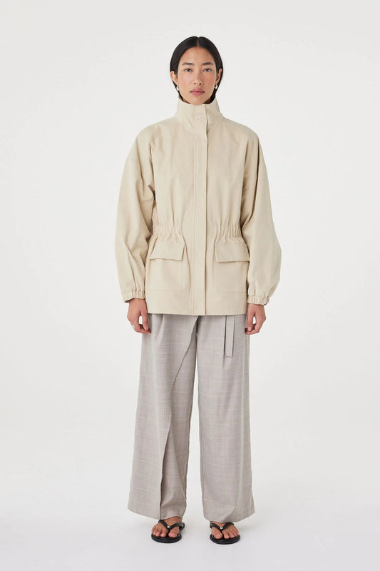 A model stands against a plain white studio background wearing a light beige high-collar jacket with front flap pockets, paired with light gray wide-leg trousers and black sandals.