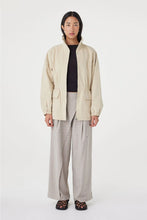 A model stands against a plain white studio background wearing a light beige jacket over a black top, paired with wide-leg light gray trousers and black patterned sandals.