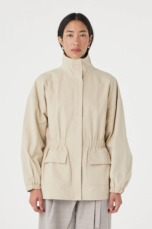 A model wearing a light beige utility jacket with a high stand-up collar, elasticized cuffs, and front flap pockets, paired with light gray trousers, against a plain light gray background.