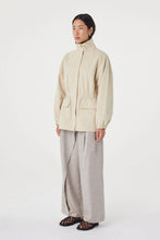 A model stands against a plain light gray studio background wearing a light beige high-neck jacket with front pockets, loose light gray wide-leg trousers, and black patterned flat sandals.