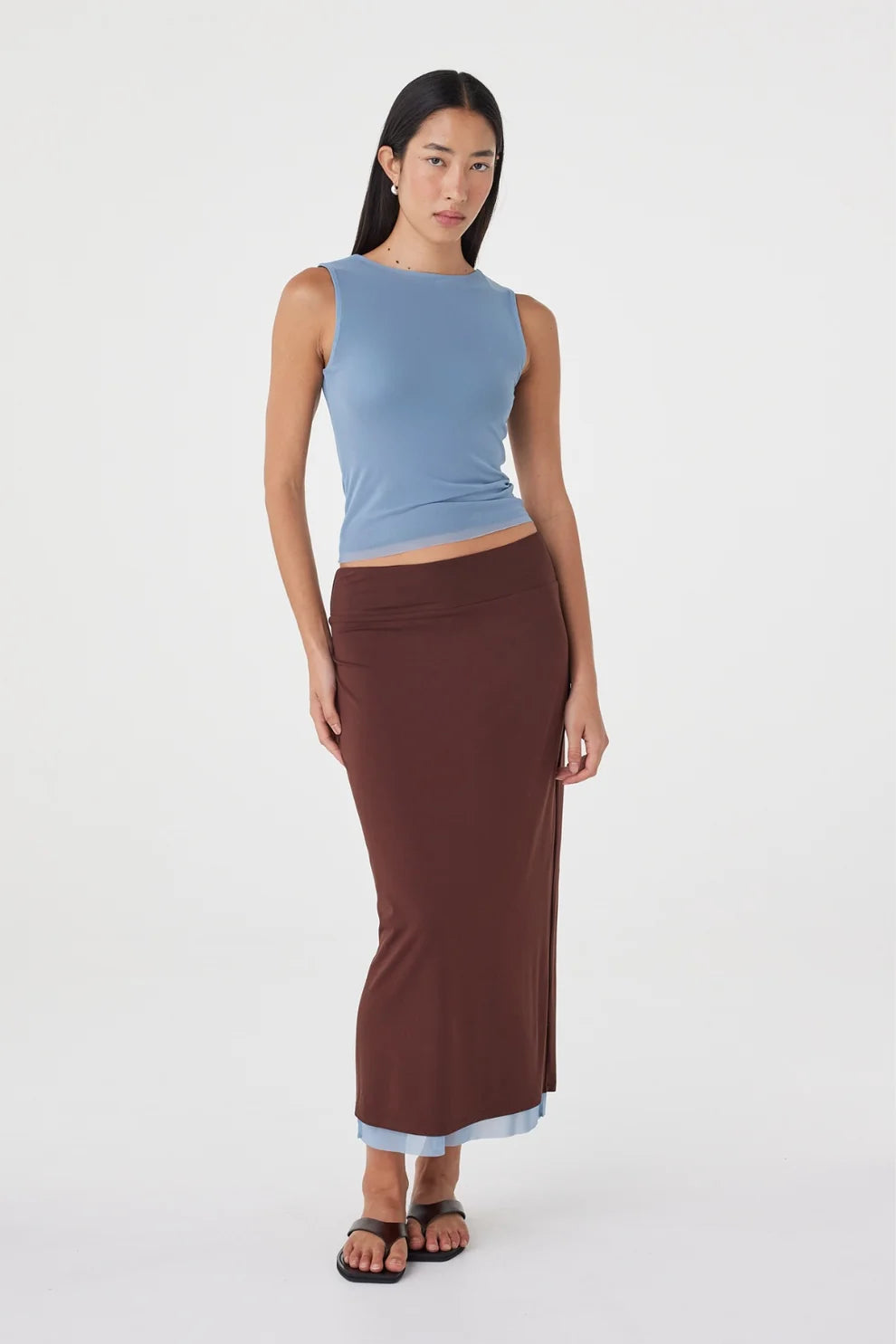 Front view of the espresso side of the reversible maxi skirt styled with a pale blue sleeveless top and black sandals against a white studio background.