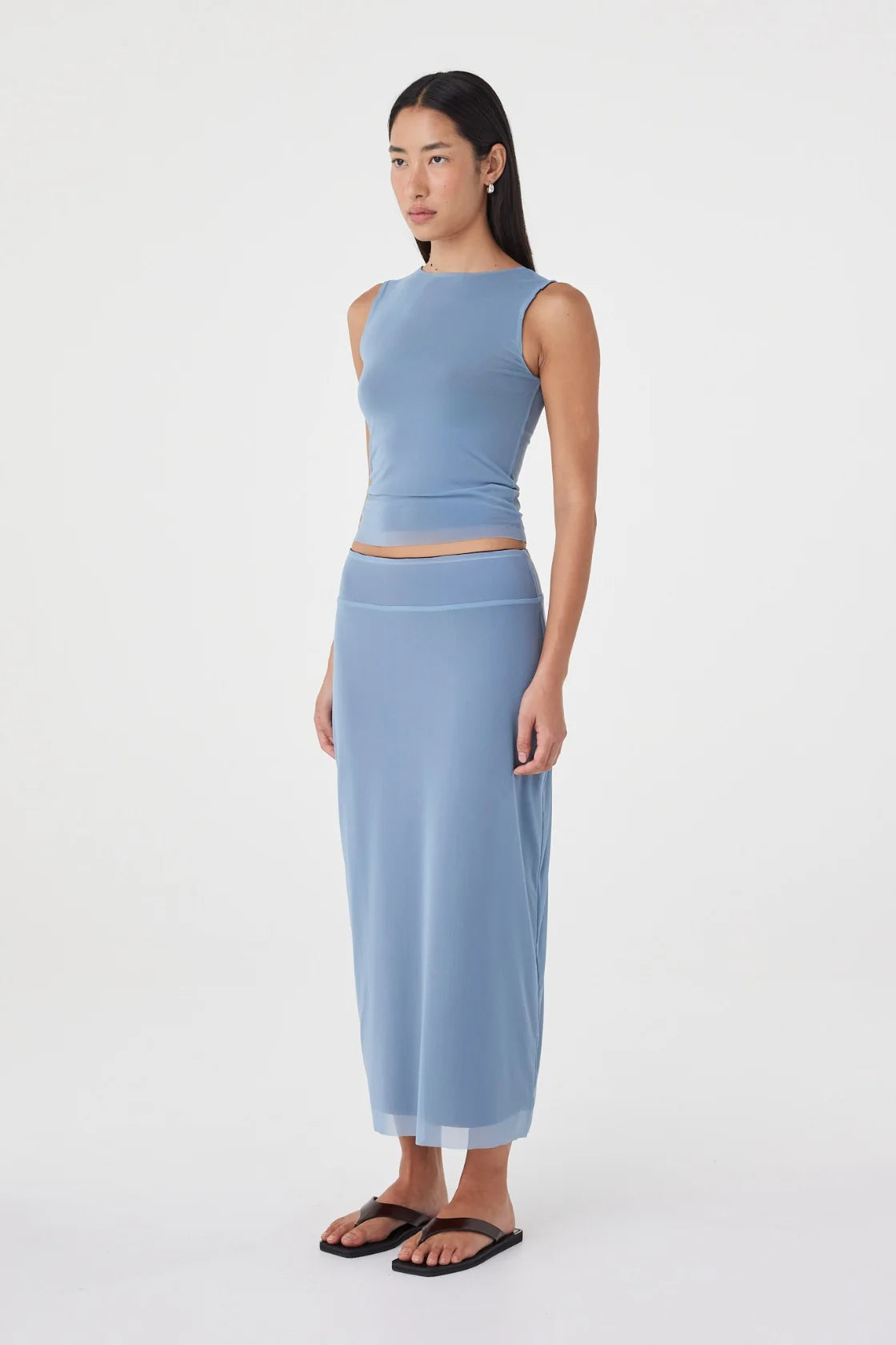 Three-quarter side view of the pale blue reversible maxi skirt styled with a matching pale blue sleeveless top and black sandals in a minimal white studio.