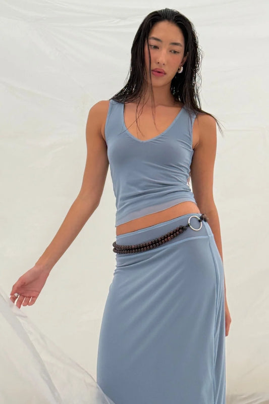 Pale blue reversible maxi skirt styled with a matching pale blue sleeveless top and beaded belt in a relaxed pose against soft white curtains.