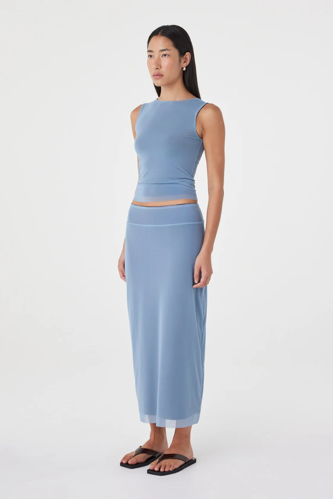 Three-quarter side view of the pale blue sleeveless fitted top paired with the matching pale blue maxi skirt and black sandals in a minimal white studio.