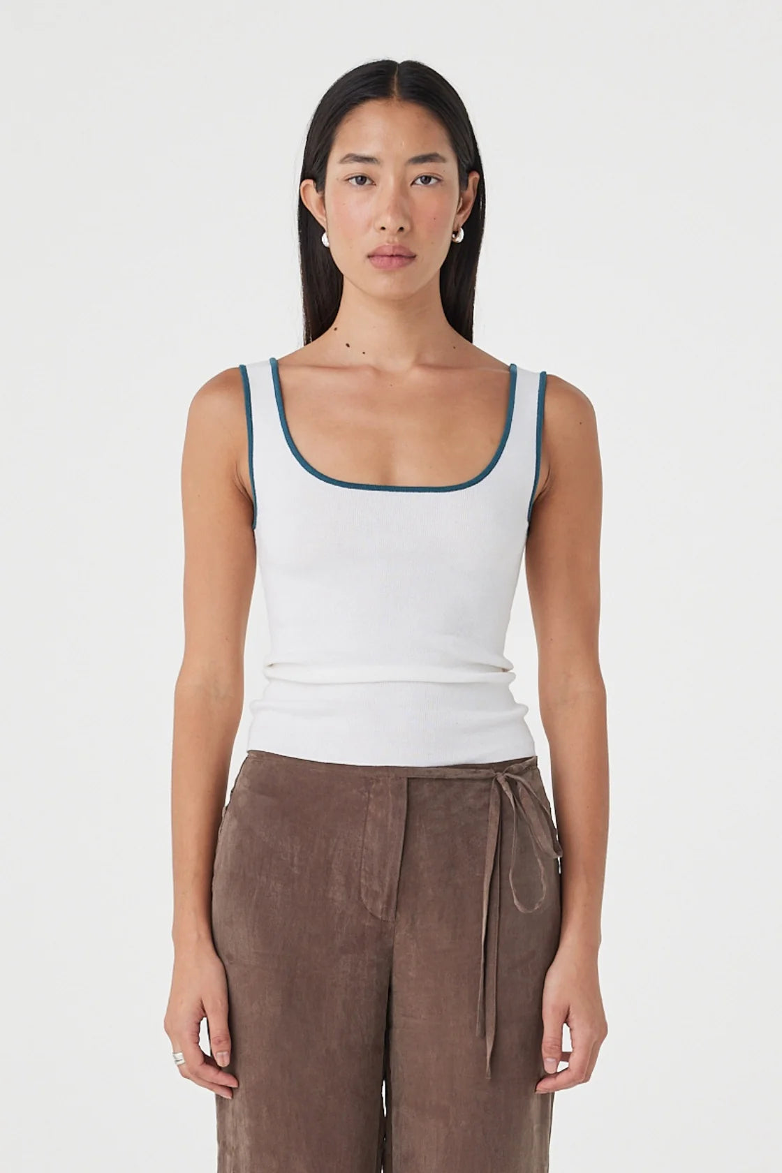 Close-up front view of the cream fitted tank showing the teal contrast trim and scoop neckline, styled with brown wide-leg wrap pants against a clean white studio background.