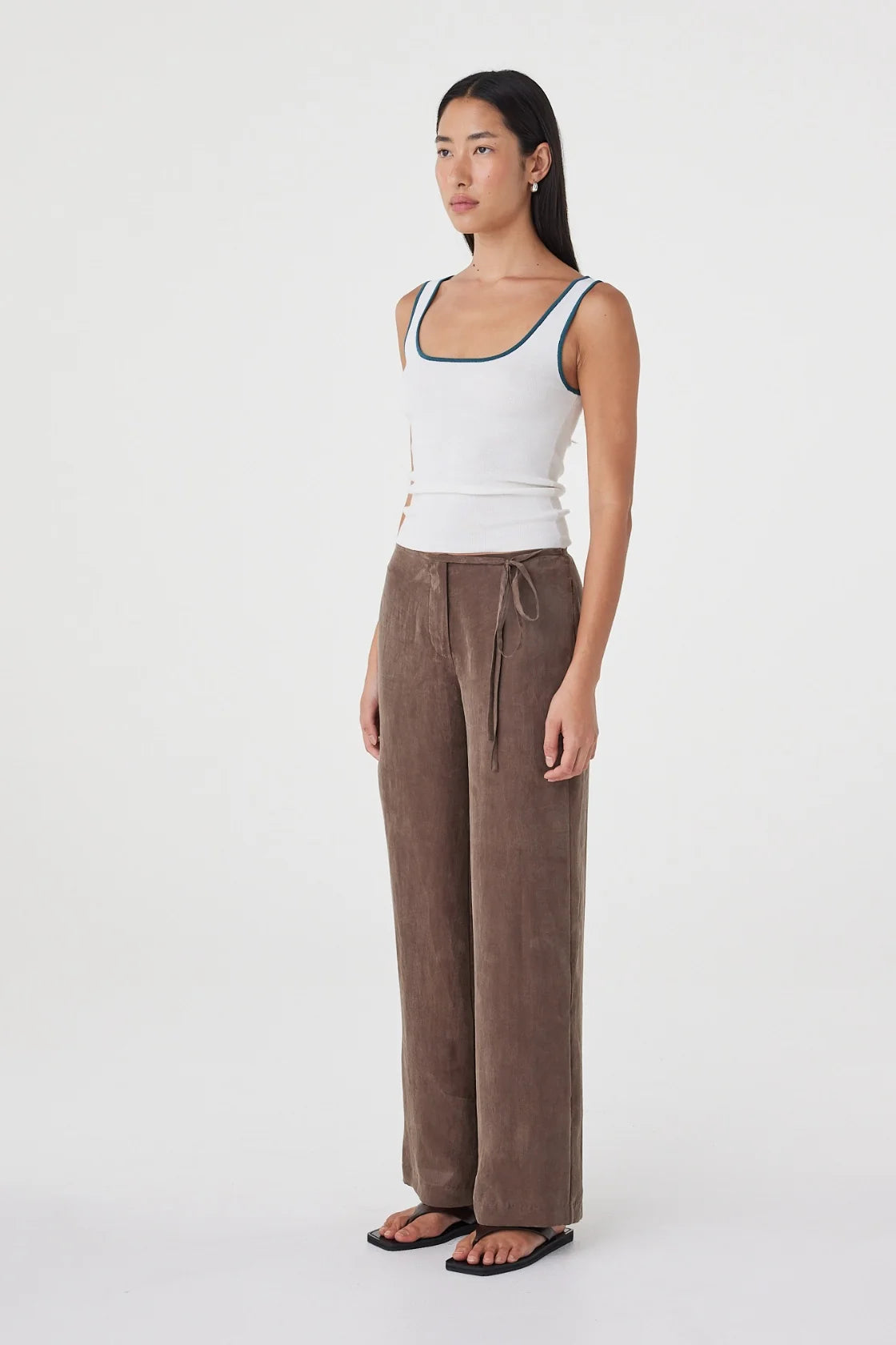 Three-quarter side view of the cream fitted tank with teal contrast trim, styled with brown wide-leg wrap pants and black sandals in a minimal white studio.