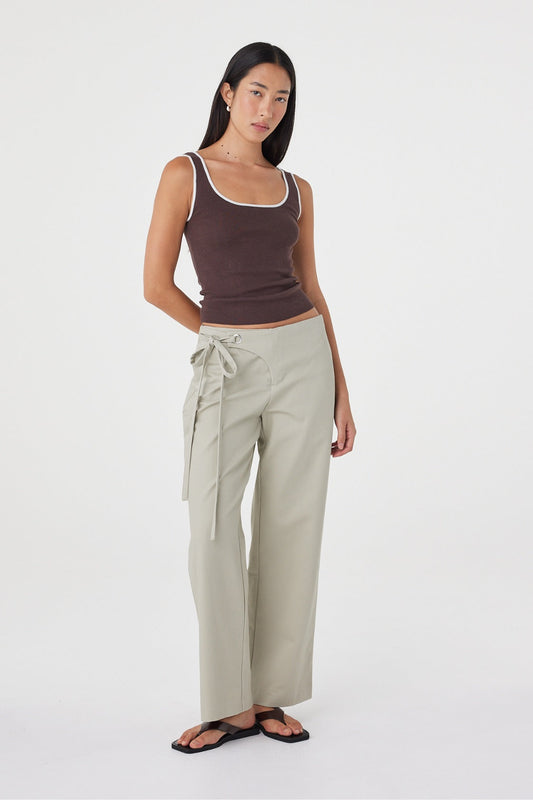 Model wearing the espresso fitted tank with cream wrap-style trousers, showing the contrast neckline and armhole trim, slim silhouette and hip-length finish in an angled front view.