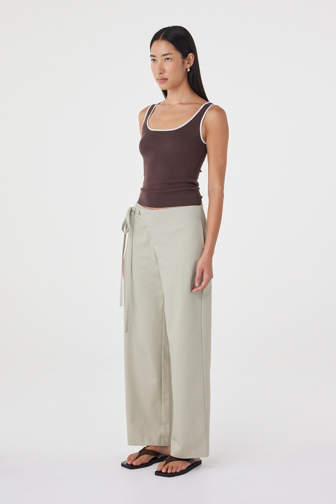 Model wearing the espresso tank with cream tailored trousers and black sandals, showing the square scoop neckline, contrast edging, fitted waist and streamlined silhouette in an angled side view.