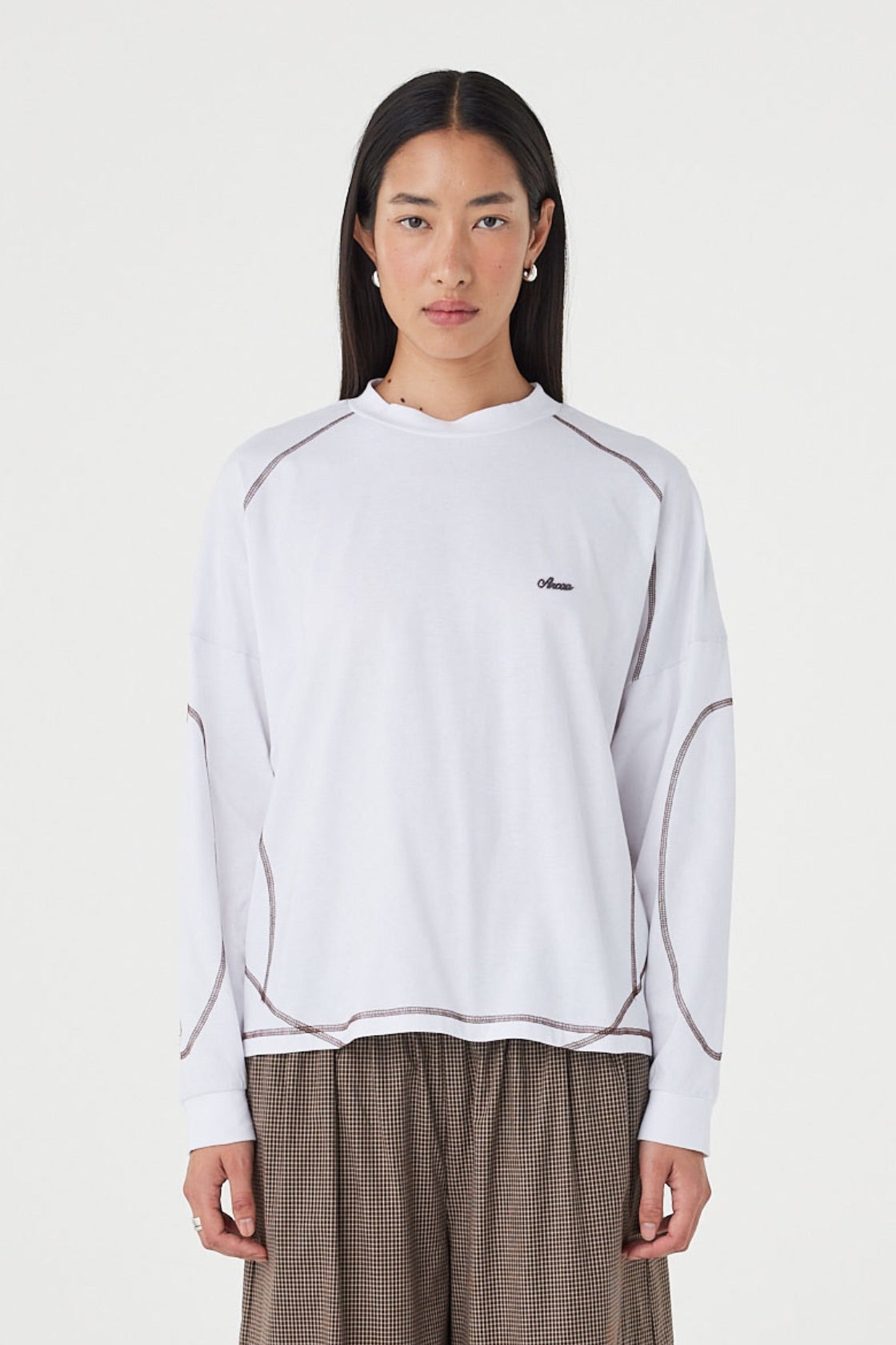 Close-up of the cream long-sleeve tee, showing the crew neckline, dropped shoulders, espresso overlock stitching, curved seam detailing and relaxed fit.