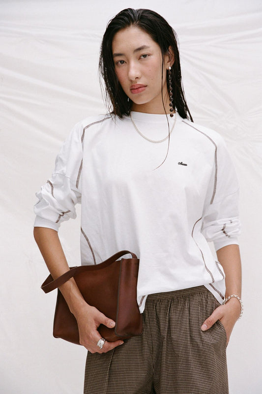 Model wearing the cream long-sleeve tee with brown check trousers and a brown handbag, showing the crew neckline, contrast seam detailing, relaxed sleeves and loose silhouette in a lifestyle close-up.
