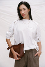 Model wearing the cream long-sleeve tee with brown check trousers and a brown handbag, showing the crew neckline, contrast seam detailing, relaxed sleeves and loose silhouette in a lifestyle close-up.